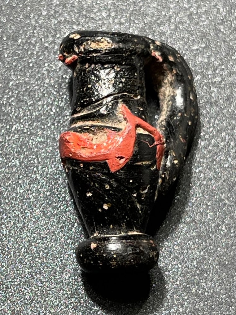 Romersk antikk Ugjennomsiktig Attraktiv amulett formet som en klassisk Oinochoe-vase med en kurvet rødfarget ornament. Med en #1.2